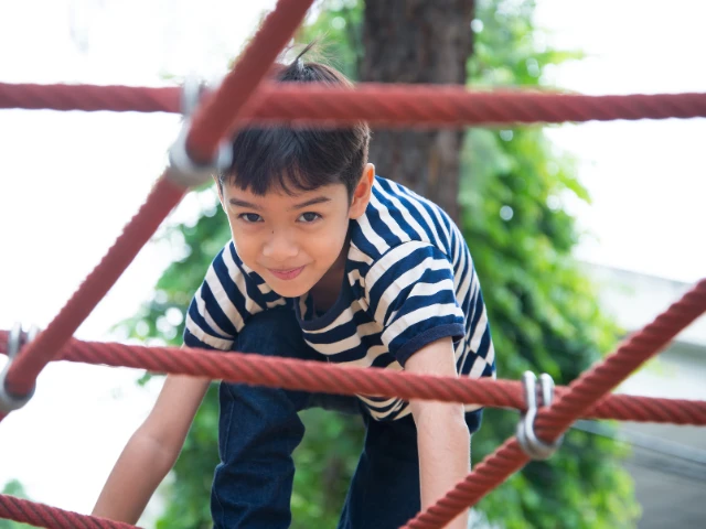 Child climbing a rope playground structure outdoors, smiling while navigating the ropes, with trees and greenery softly blurred in the background.