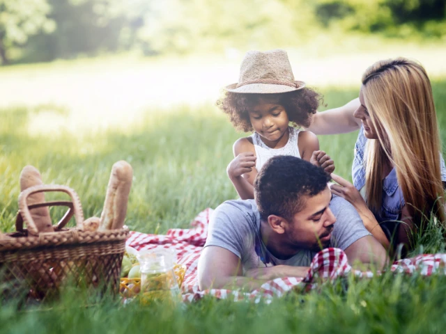 Family enjoying a picnic on a blanket in a grassy park, with parents and a young child relaxing together beside a picnic basket on a sunny day.