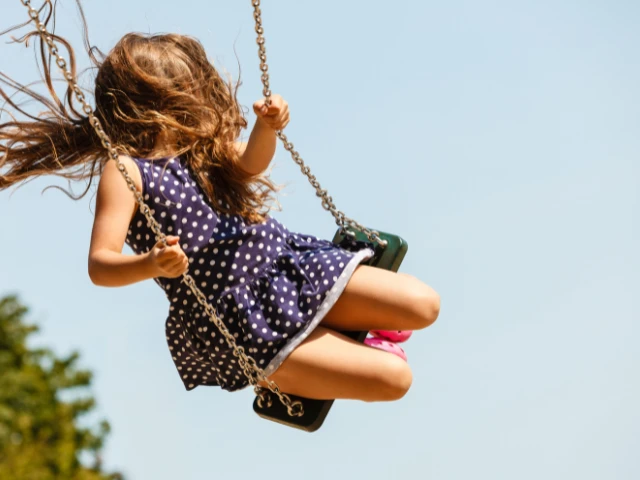 Child swinging high on a playground swing, with hair blowing in the breeze against a bright blue sky, capturing a fun and carefree outdoor moment.