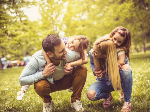 Family enjoying a playful day outdoors, with two parents giving their young daughters piggyback rides in a sunny, tree-filled park.
