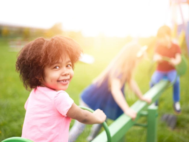Young child smiling while playing on a seesaw at a sunny outdoor playground, with other children playing in the background.