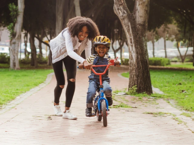 Parent helping a smiling child learn to ride a bicycle along a tree-lined park path.