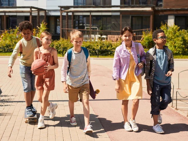 Group of school-age children walking together outdoors with backpacks, a basketball, and skateboards.