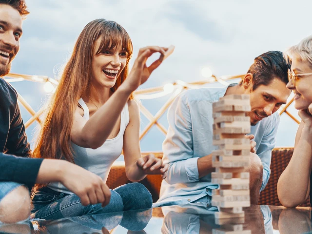 Group of friends laughing and playing a wooden block-stacking game together outdoors.