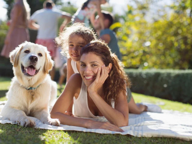 Smiling mother and child relaxing on a blanket with their dog in a sunny outdoor park.