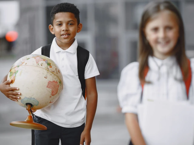 Student holding a globe and wearing a backpack outside a school, with another student in the background.
