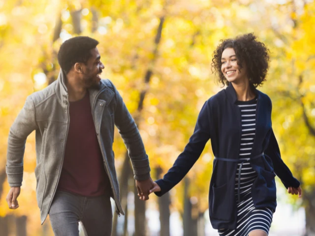Smiling couple holding hands while walking together through a tree-lined park in autumn.