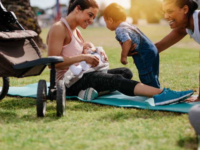 Two women relaxing outdoors with a baby and young child beside a stroller on a sunny day.