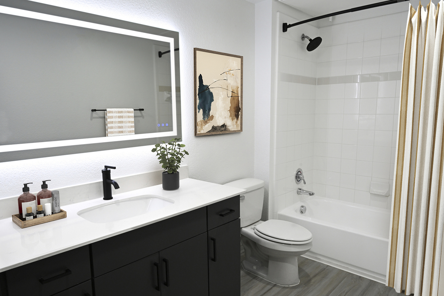 Modern bathroom featuring sleek black fixtures, a large illuminated mirror, elegant countertop with toiletries, and a chic abstract painting.