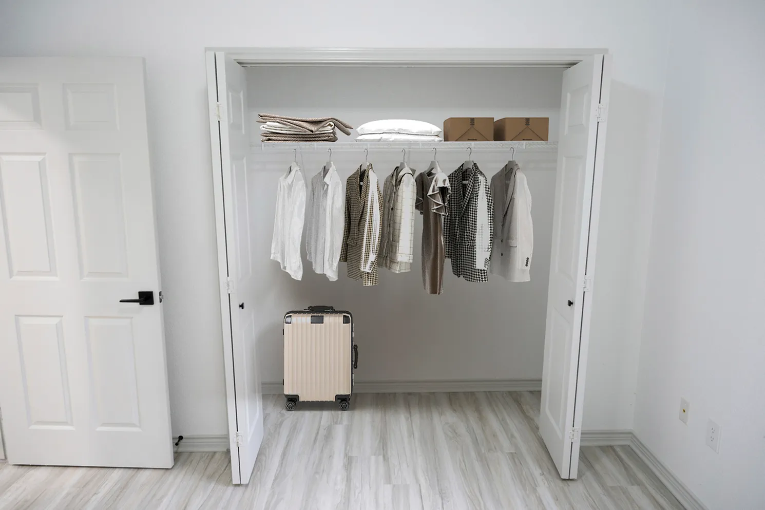 Contemporary apartment closet featuring organized clothing storage with sleek hangers, folded blankets, and a stylish suitcase on polished wood floors.