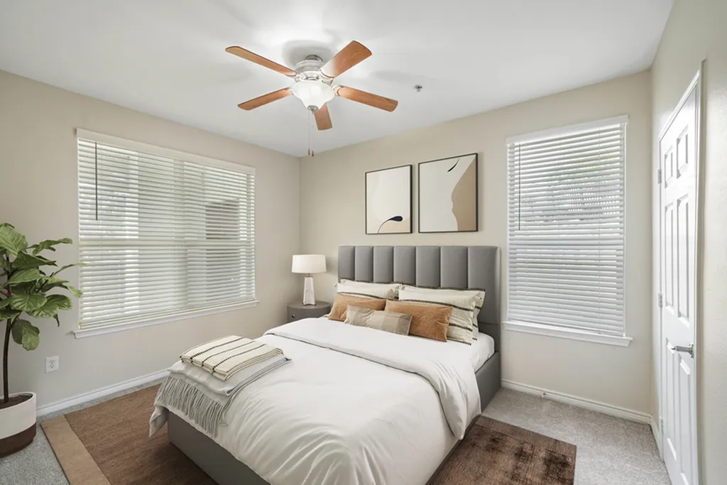 Bedroom with plush carpeting, ceiling fan, large window, plotted standing plant, queen size bed with white bedding, two large windows, window art