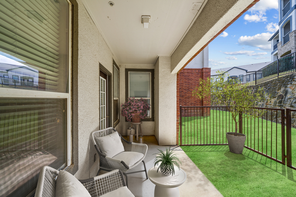 Patio with lush green grass, black railing, patio chairs with pillow, and potted plants.