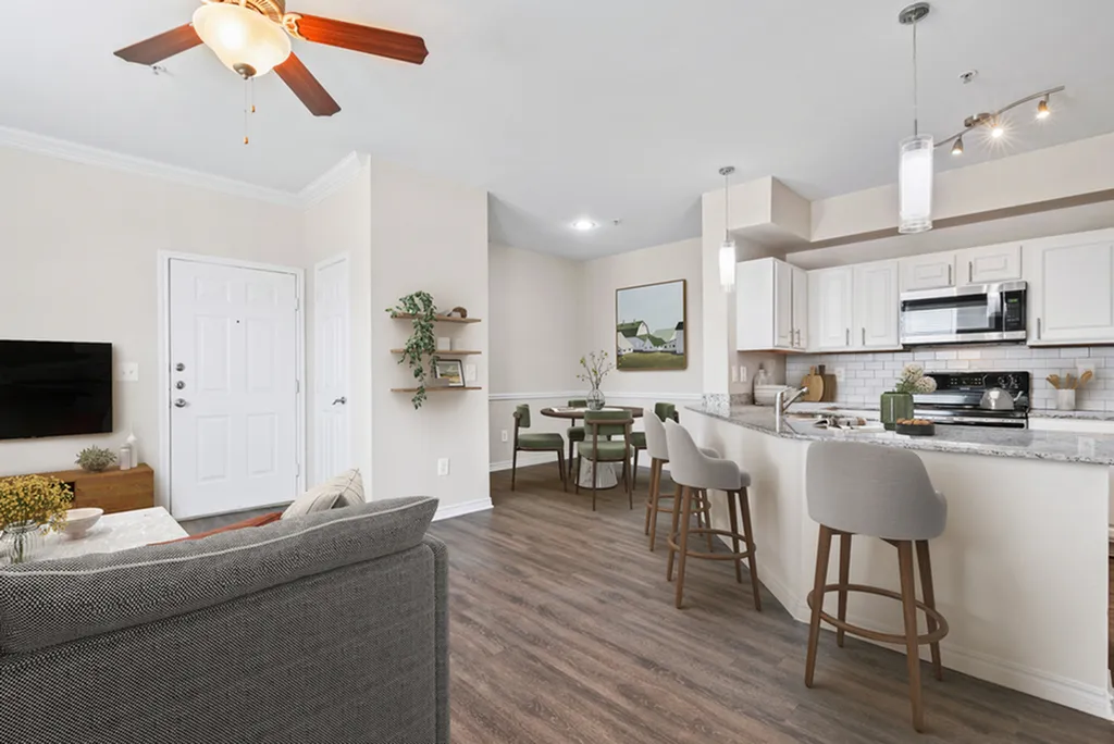Kitchen with pendant lighting, granite counters, stainless steel appliances, white tile backsplash, hardwood flooring, white cabinetry, overlooking dining area and livingroom with ceiling fan, grey couch, and television