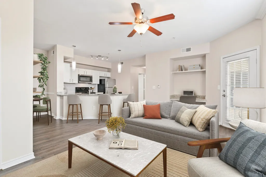 Living room with ceiling fan, wooden floors with a yellow rug, coffee table, grey couch, beige chair, with built-in shelving and desk area