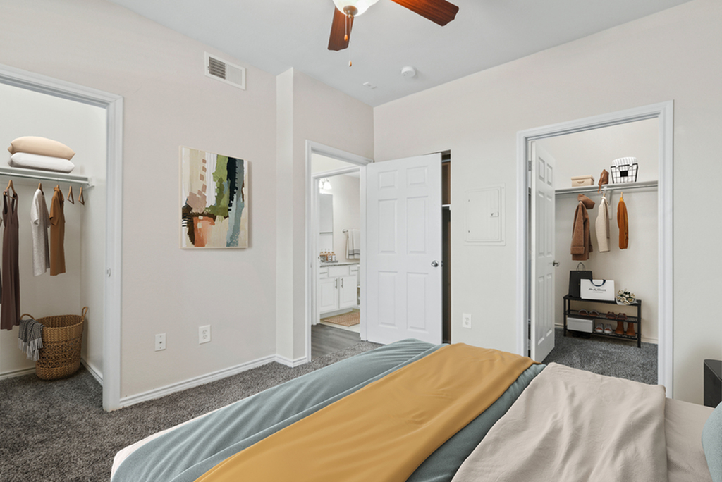 Bedroom with ceiling fan, bed with blue and brown bedding, plush grey carpeting, wall art, and two separate closets