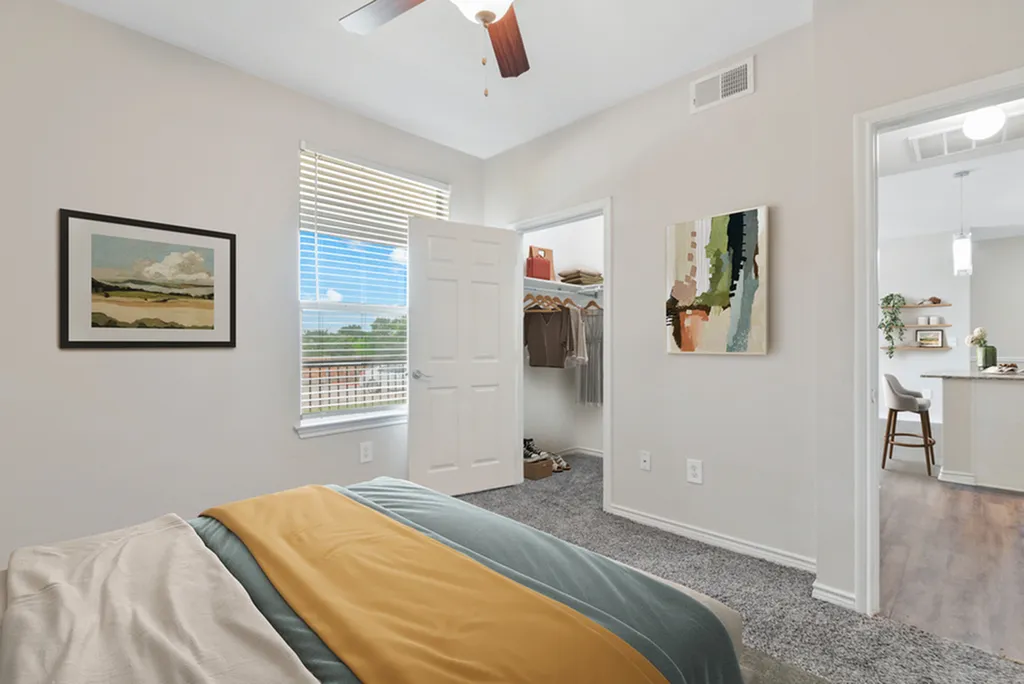 Bedroom with ceiling fan, bed with blue and brown bedding, plush grey carpeting, wall art, large window, open closet door with clothe, open bedroom door looking into living area