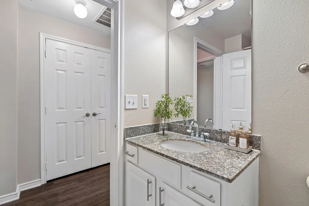 Bathroom with granite counters, large vanity mirror with lighting, hardwood floors, white cabinetry, and decorative plant