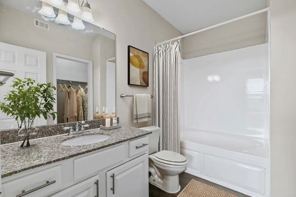 Bathroom with large vanity mirror and lighting, granite counter tops, toilet, wall art, towel rack with tan towel, bathtub and shower and shower curtains