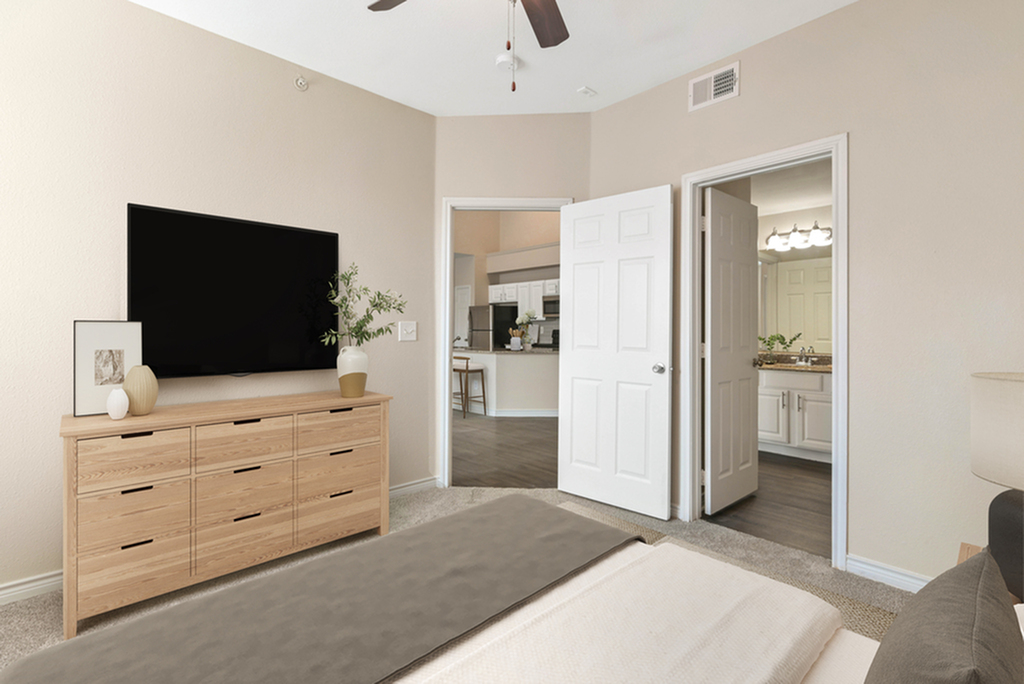 Bedroom with ceiling fan, large bed with tan and white bedding, wooden dresser with wall mounted TV open door looking into bathroom, and open door looking into living room