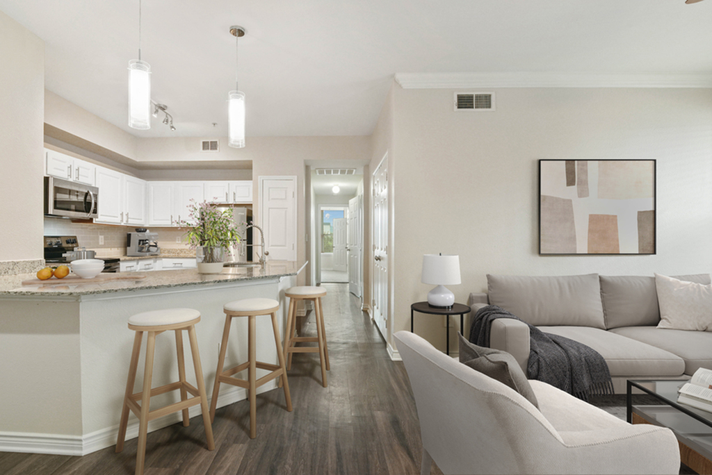 Open Concept living room with kitchen pendant lighting, granite counters, bar stools, white cabinetry, grey couch and chair, hardwood style flooring, and wall art.