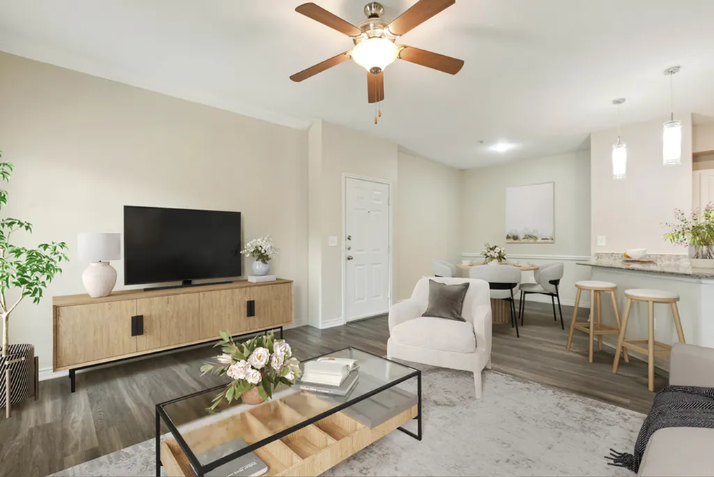Open-concept living room with glass coffee table, hardwood flooring and grey rug, ceiling fan, wooded entertainment stand and TV, leading to a  dining area and breakfast bar with pendant lighting.