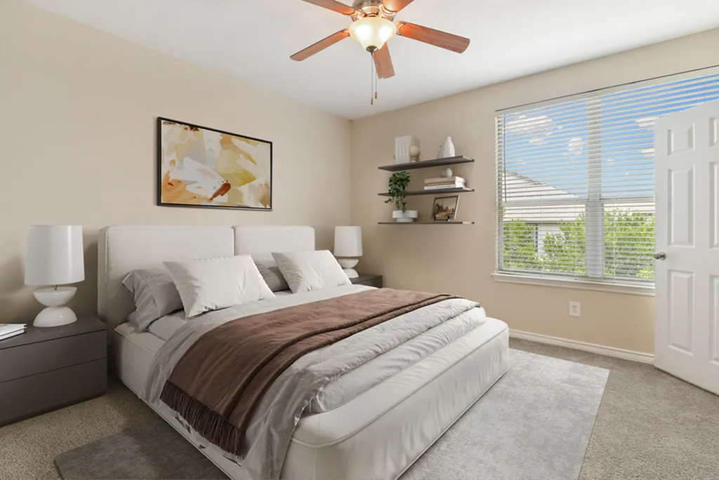 Bedroom with ceiling fan, king size bed with cream beddings, wall art, plush carpeting, nightstand with white lamp, wall shelving, large window.