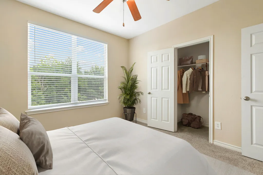 Bedroom with ceiling fan, king size bed with cream beddings, wall art, plush carpeting, large window. closet door opened showcasing clothes