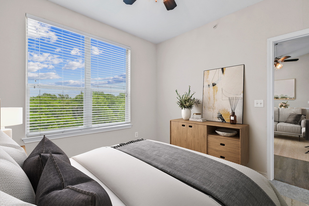 Bedroom with ceiling fan, large bed with cream and gey bedding, wooden dresser with potted plant and wall art, a large window and the bedroom door open looking into the living room