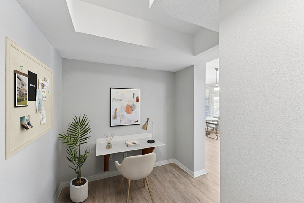 Entryway nook with desk, white chair, bulletin board, large potted plant, and wall art