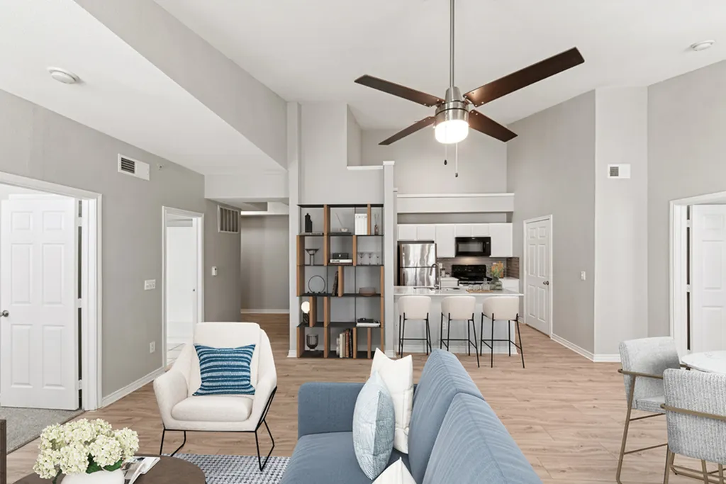 Living room with ceiling fan, hard wood flooring, blue rug, blue couch, and white chair, intricate bookshelf, looking into kitchen with pendant lighting, stainless steel appliances, and creme bar chairs