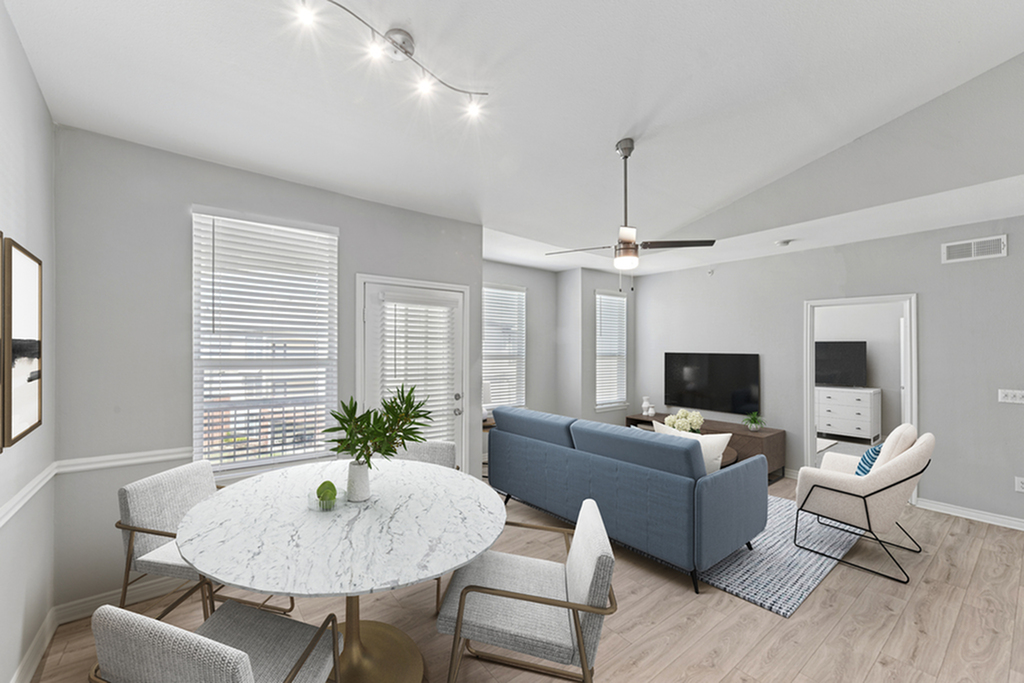 Open concept living and dining room with ceiling fan, round dining table and three cream chairs, blue couch, white cushioned chair, wall mounted flat screen, ample windows for natural lighting