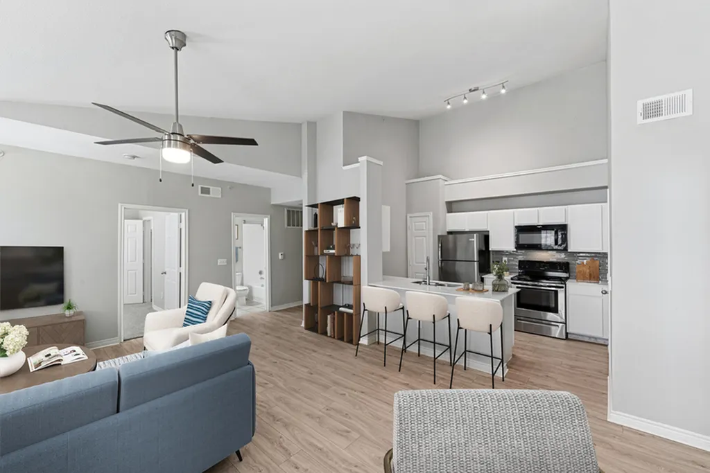 Living room with ceiling fan, hard wood flooring, blue rug, blue couch, and white chair, intricate bookshelf, looking into kitchen with pendant lighting, stainless steel appliances, and creme bar chairs