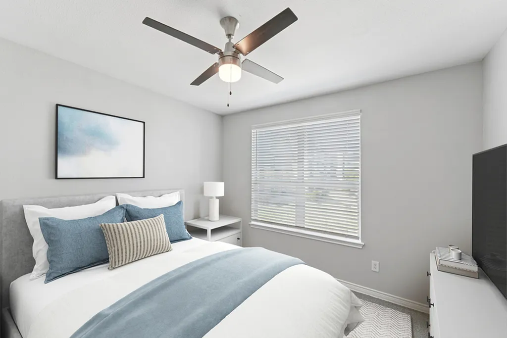 Primary bedroom with ceiling fan, large bed with blue and white bedding, grey night stand with lamp, and wall art, flat screen TV, and large window