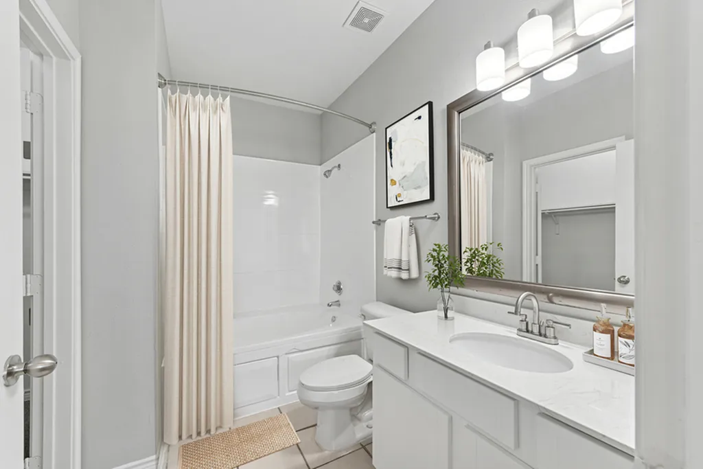 Primary bathroom with large vanity mirror and vanity lighting, quartz counters, toilet, tan rug, towel rack and white towel, wall art, shower and tub with curved shower rod and cream colored shower curtain