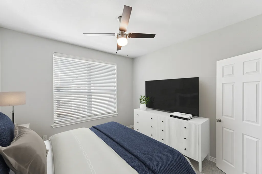 Secondary bedroom with ceiling fan, large bed with white and blue bedding, white dresser with TV and potted plants, large window, and lamp