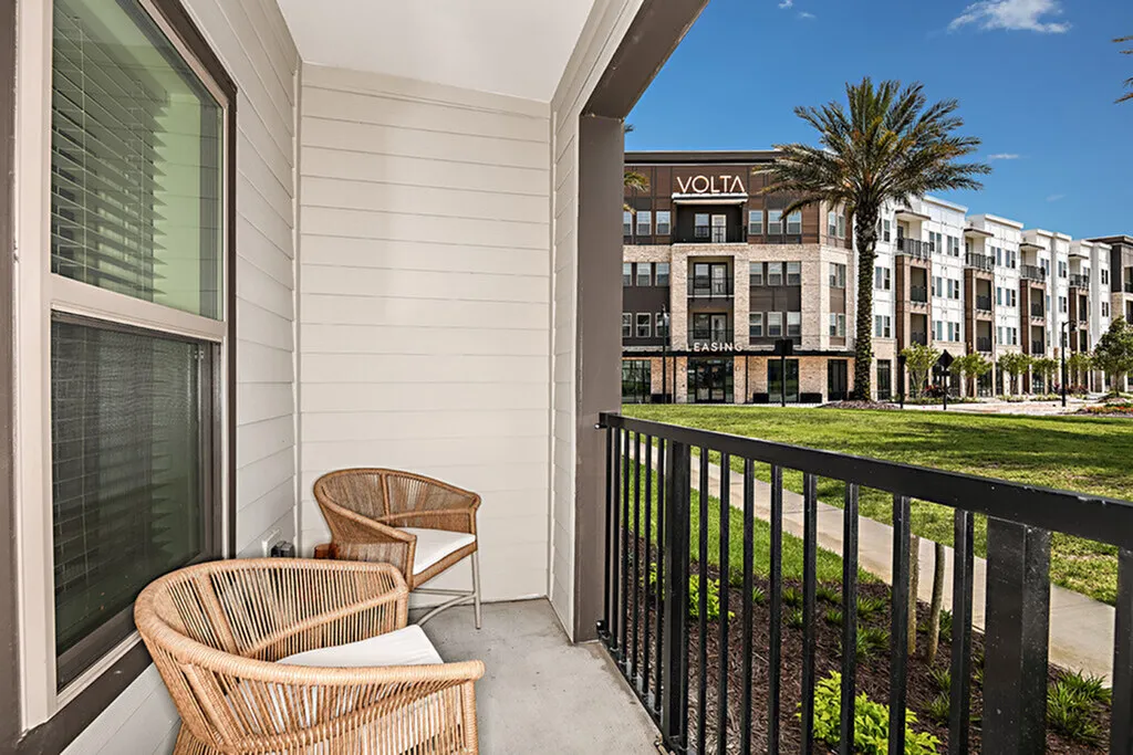 Charming apartment balcony with cozy rattan chairs and a view of modern apartment buildings and lush greenery.