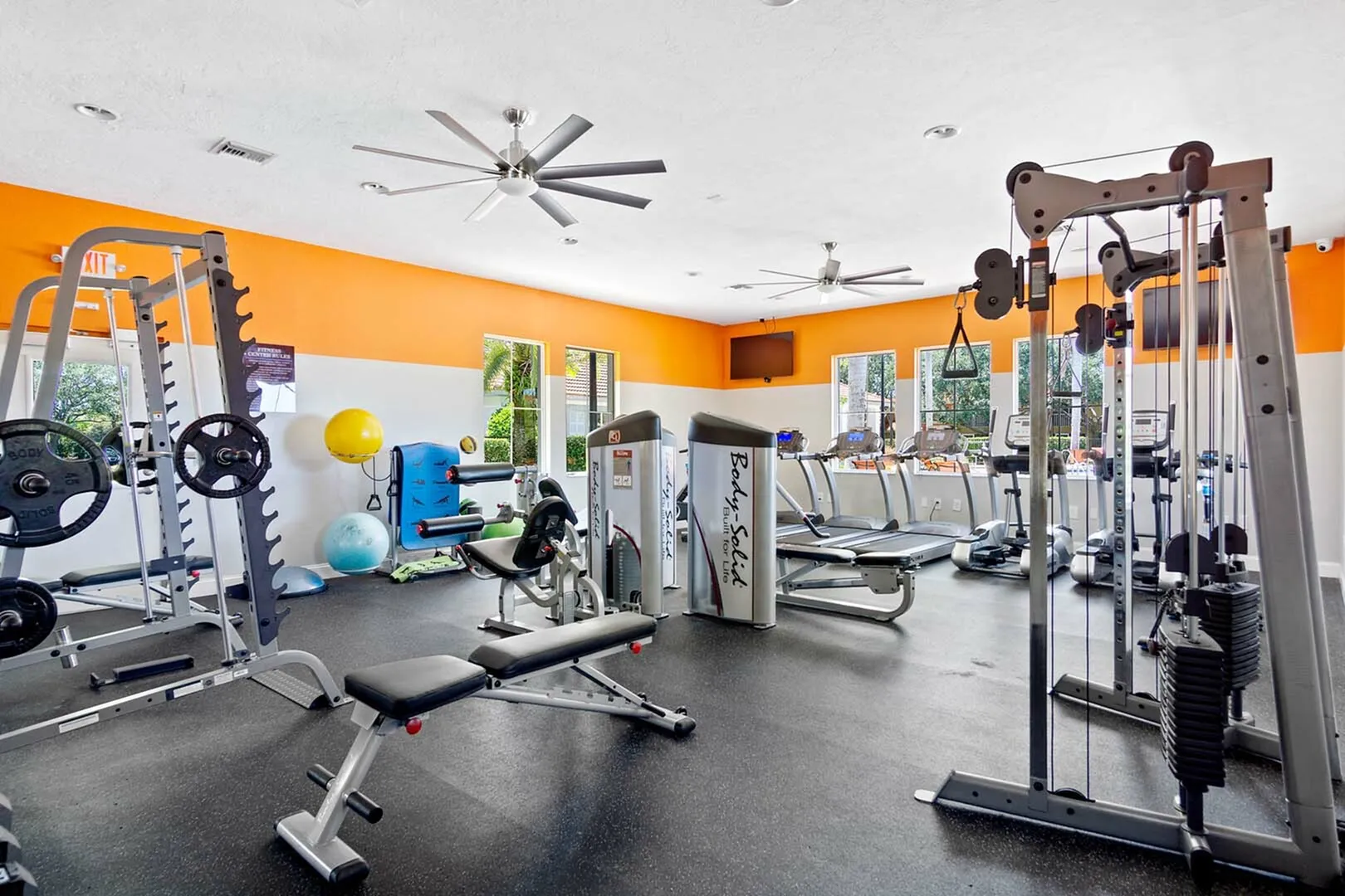 Fitness Center with cardio and strength stations, large fans and tv mounted on wall.