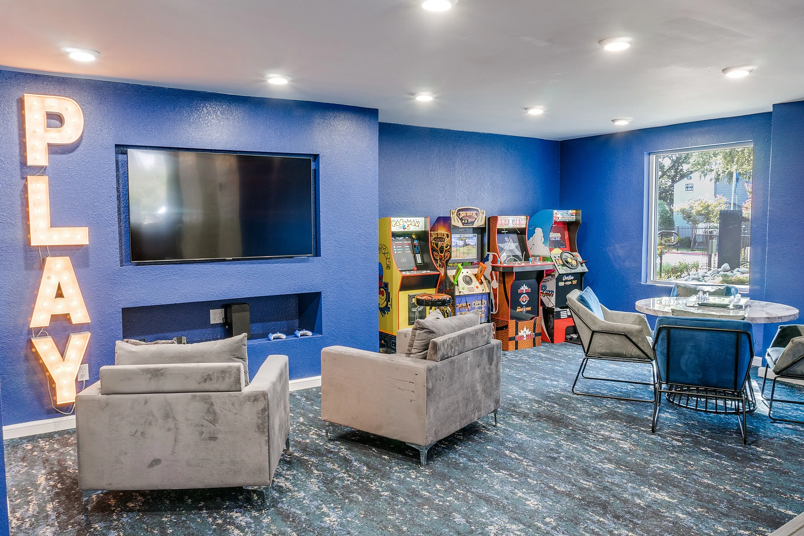Bright game room with blue walls featuring cozy seating, retro arcade machines, and a large wall-mounted TV.