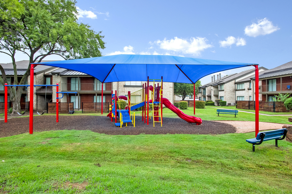 Inviting apartment community with a vibrant playground under a blue canopy, surrounded by lush greenery and modern buildings.