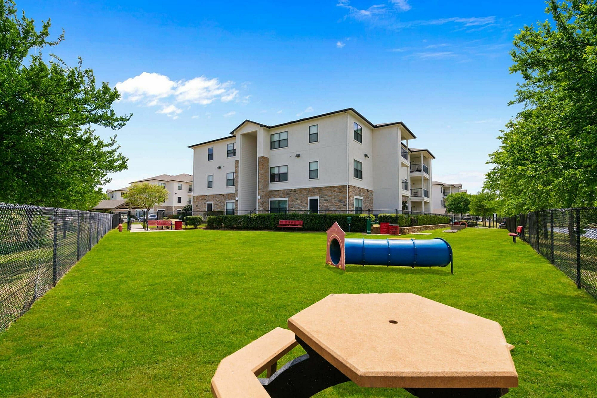 Inviting apartment building with a lush green dog park, play tunnel, and picnic area under a bright blue sky.