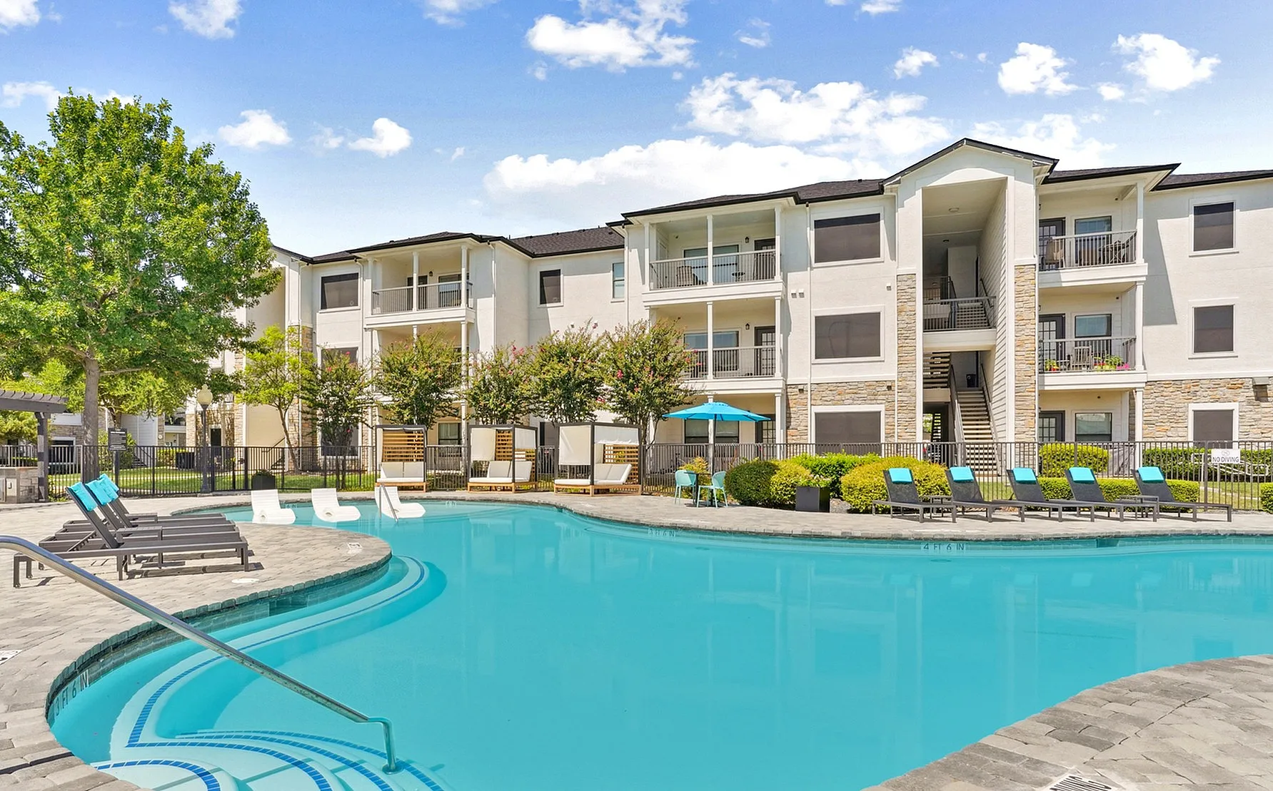 Vibrant pool area with modern lounge chairs and umbrellas, framed by a stylish three-story apartment building under a clear blue sky.