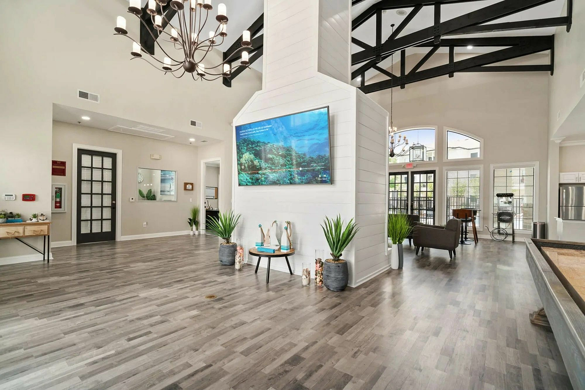 Elegant apartment lobby with high ceilings, modern chandelier, and stylish wood flooring. Features a large screen, lush plants, and ample seating, creating an inviting atmosphere.