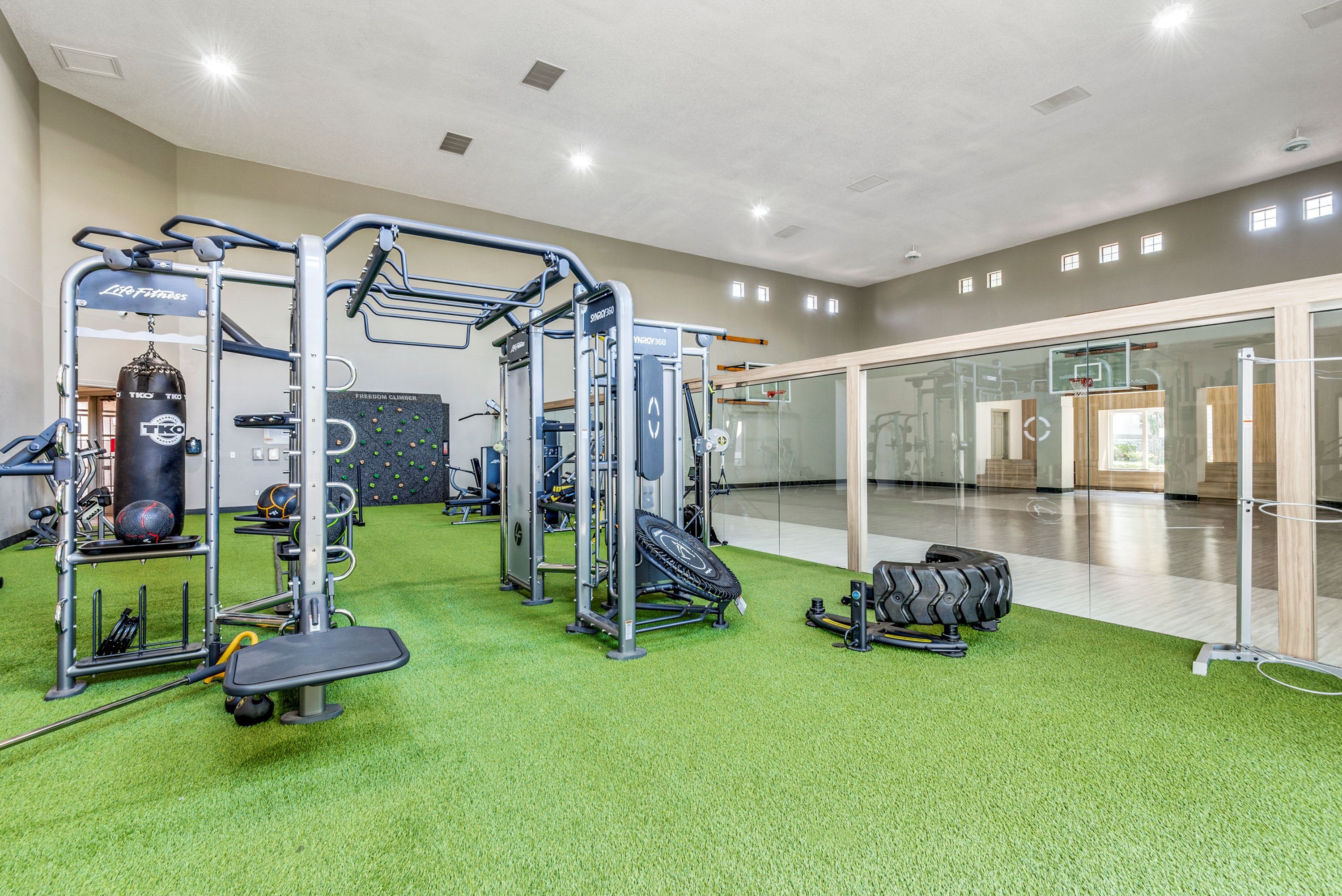 Modern fitness center boasting state-of-the-art equipment, vibrant green flooring, and a spacious indoor basketball court beyond glass walls.