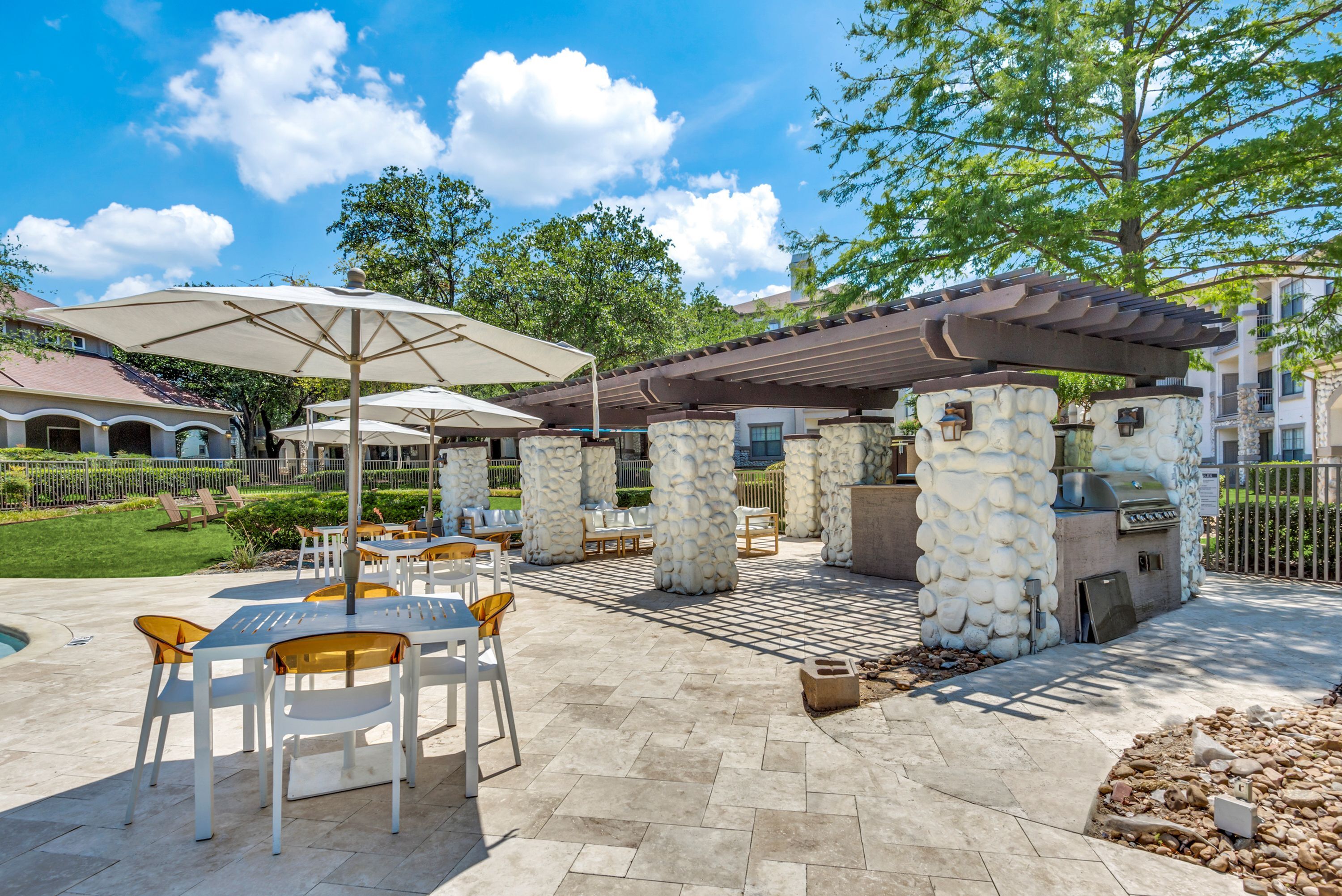 Charming outdoor patio area with stylish seating, umbrellas, and barbecue space under a clear blue sky, nestled in lush greenery.