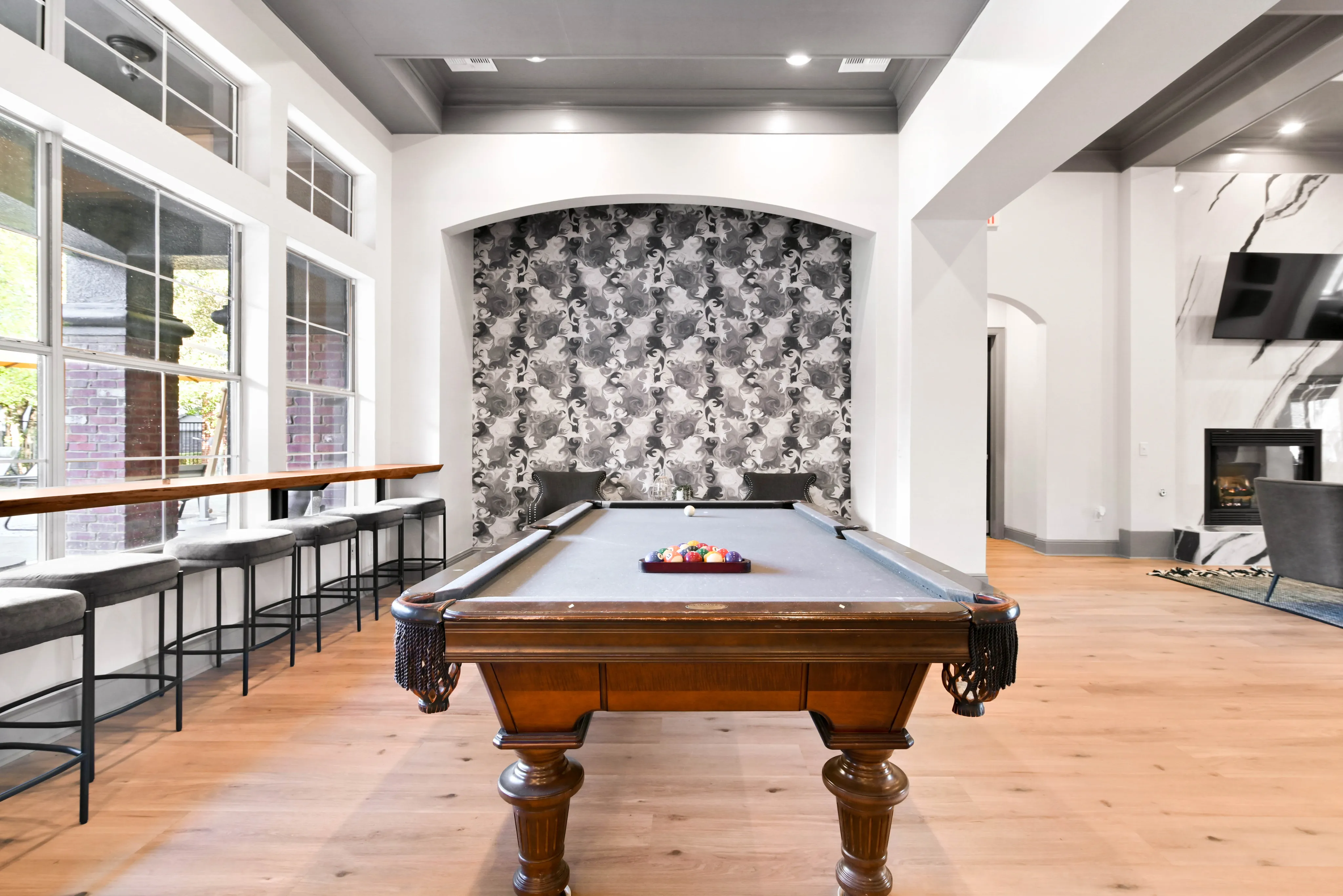 Elegant game room with a stylish pool table, modern wallpaper, and daylight streaming through large windows, perfect for entertaining.