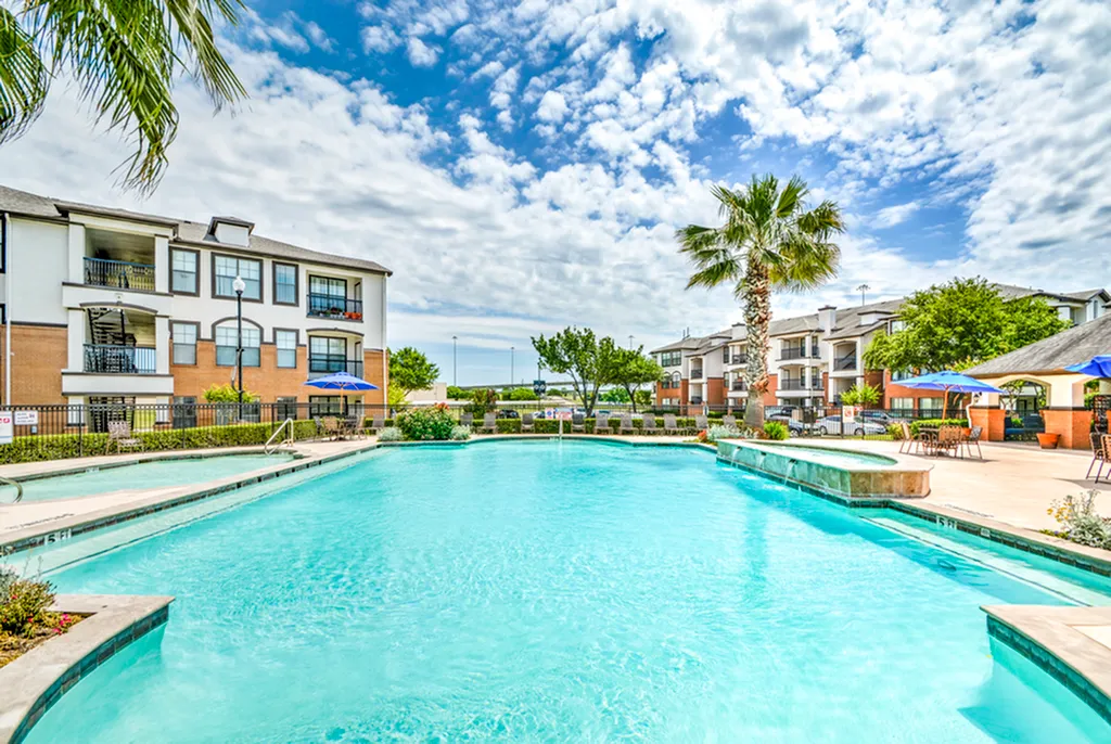 Luxurious apartment complex featuring a sparkling pool, surrounded by lush palms and stylish lounging areas, perfect for relaxing under the clear blue sky.