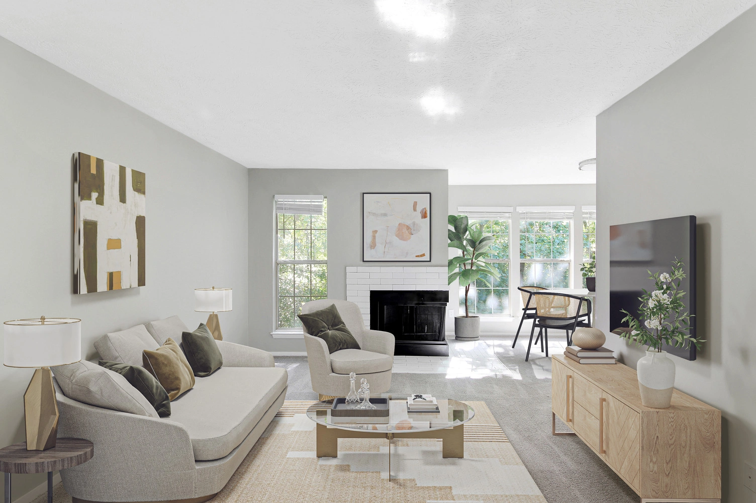 Bright, modern living room with a neutral color palette, featuring a light gray sofa, armchair, glass coffee table, and wooden media console with a mounted TV. A white fireplace sits between large windows, with natural light filling the space and a small desk area near the windows.