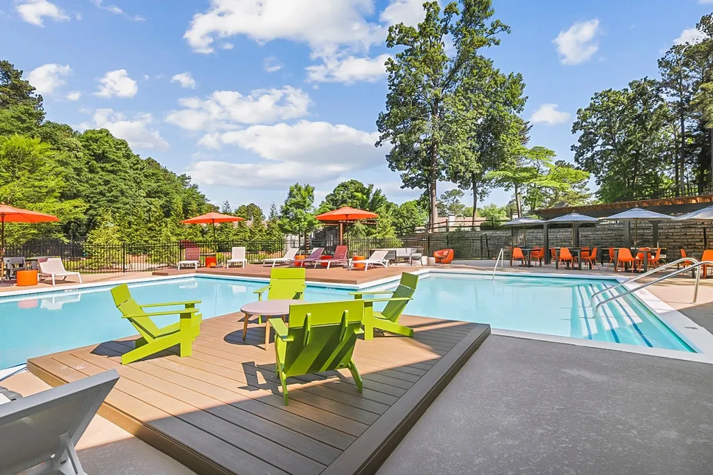 Swimming pool with expansive pool deck, bright green Adirondack chairs on a wooden deck platform, lounge seating with orange umbrellas, and a shaded dining area surrounded by trees under a blue sky.