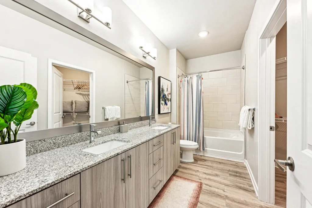 Spacious bathroom with a double-sink vanity, granite countertops, large mirror, and wood-style cabinetry, leading to a tub-shower combo with a curtain and adjacent walk-in closet.