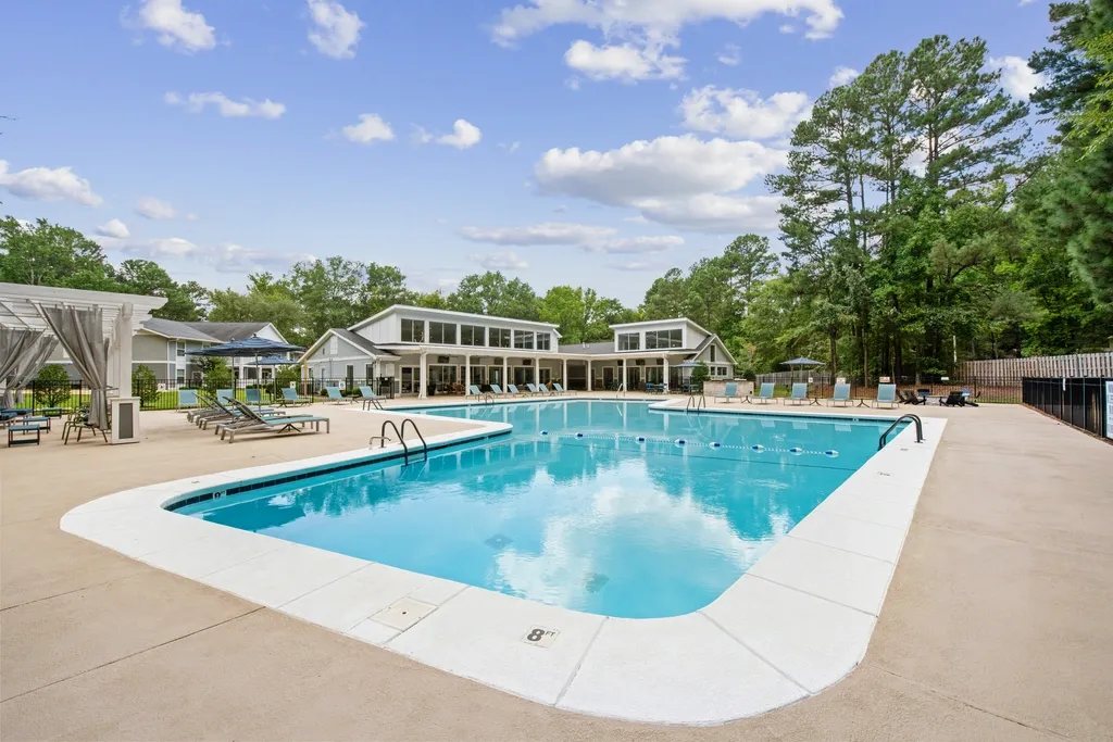 Large outdoor swimming pool with surrounding lounge chairs and umbrellas, set in front of a modern clubhouse and bordered by trees.